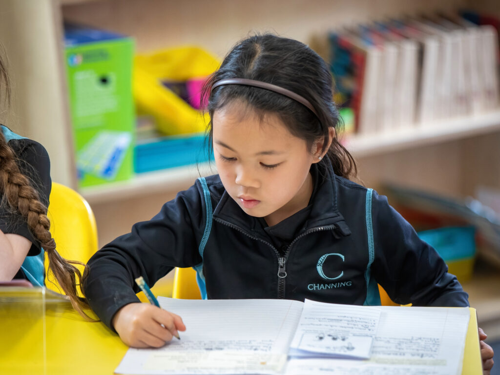 A young girl wearing a headband writes in a notebook at a bright yellow school desk.