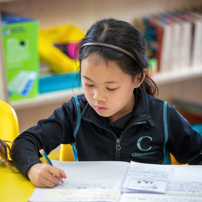 A young girl wearing a headband writes in a notebook at a bright yellow school desk.