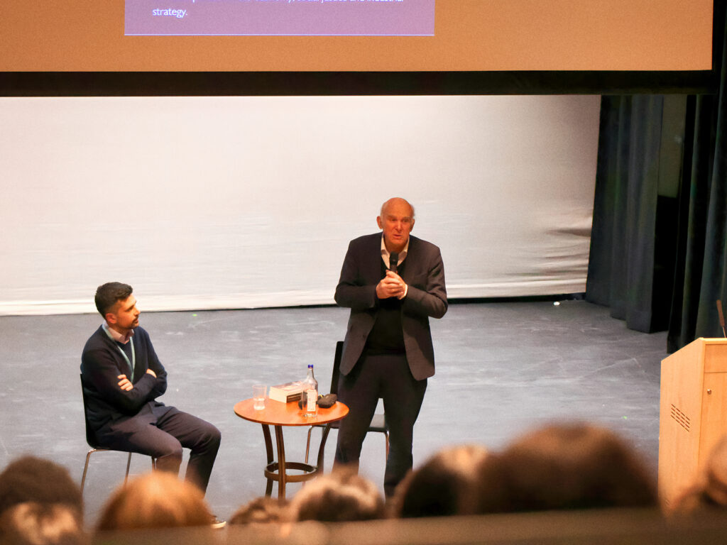 Panel discussion on a stage with two men in suits; one standing and speaking into a microphone, the other seated at a small table with water bottles, audience in foreground.