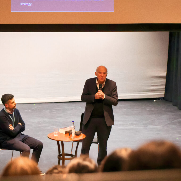 Panel discussion on a stage with two men in suits; one standing and speaking into a microphone, the other seated at a small table with water bottles, audience in foreground.
