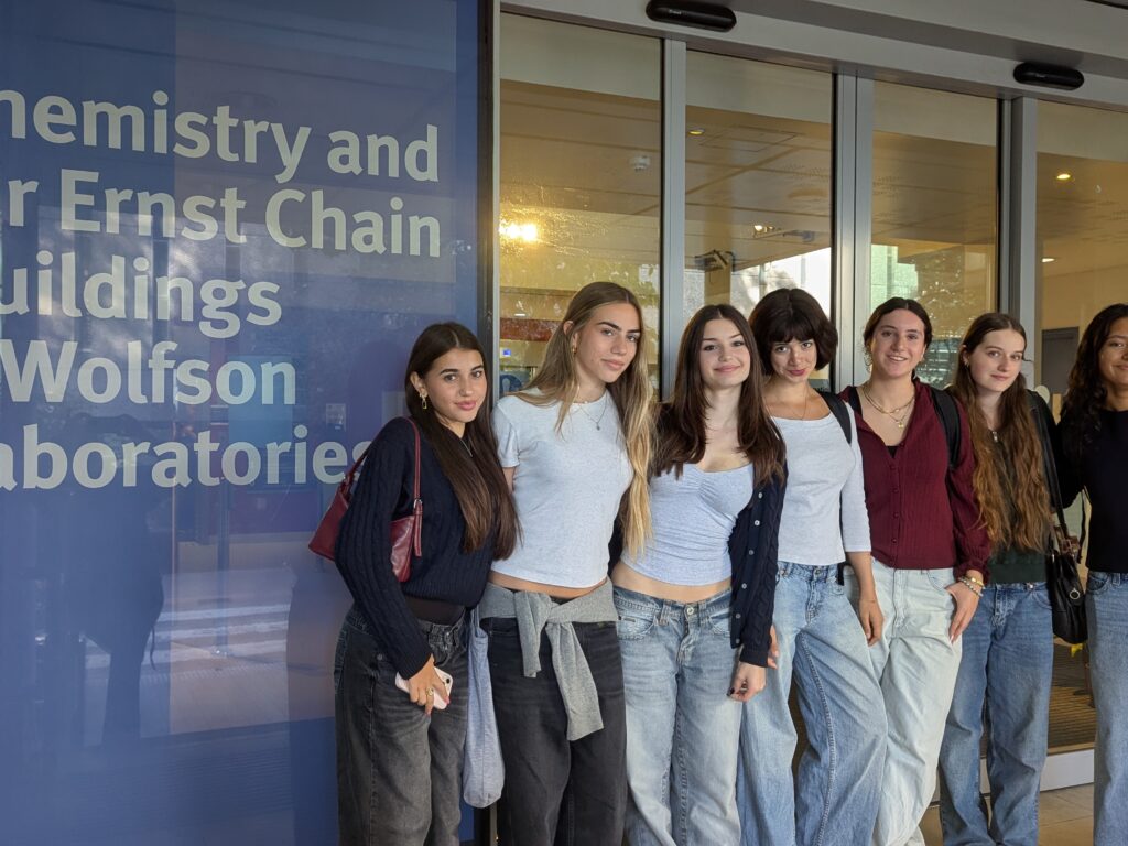 Group of seven young women posing together outside a building with a blue panel reading Chemistry and Wolfson Laboratories; students smiling at the camera, casual clothing.