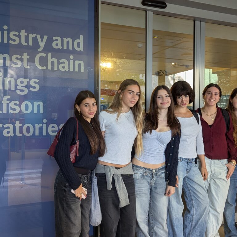 Group of seven young women posing together outside a building with a blue panel reading Chemistry and Wolfson Laboratories; students smiling at the camera, casual clothing.