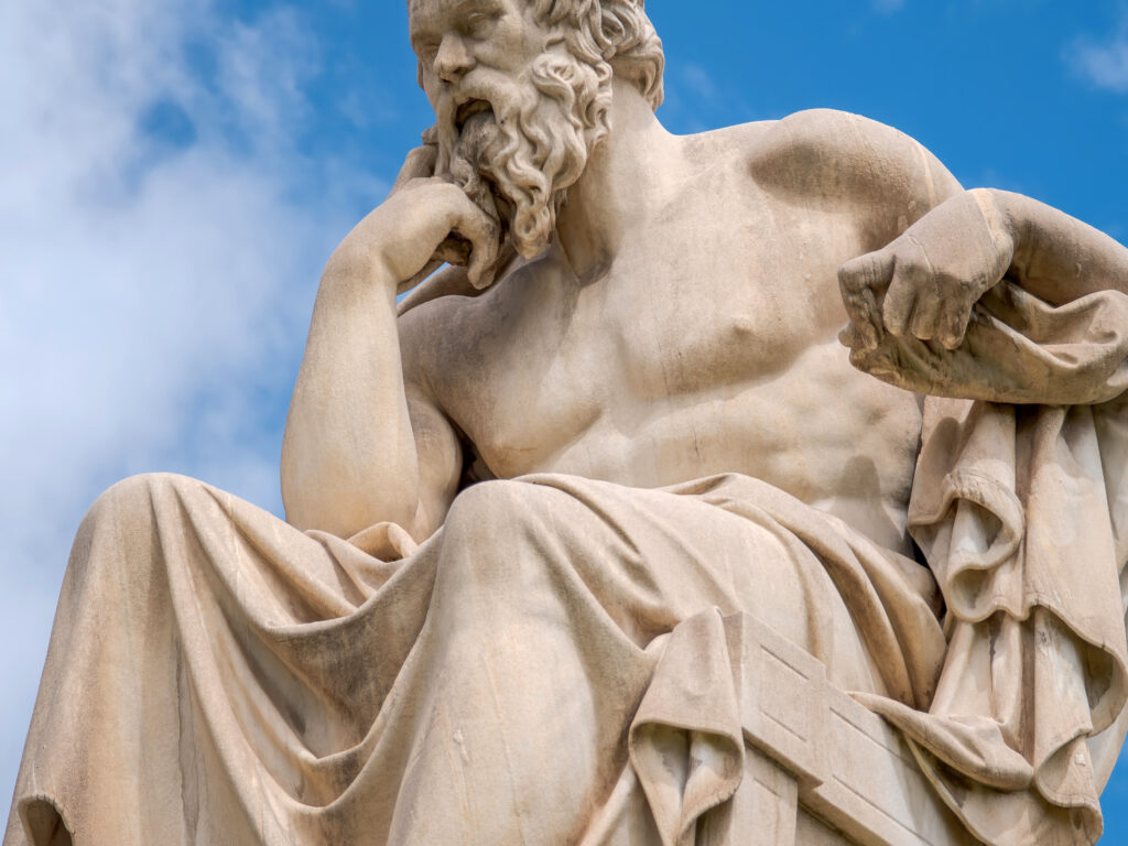 Close-up of a marble statue of a bearded male figure against a blue sky, highlighting the flowing hair and drapery