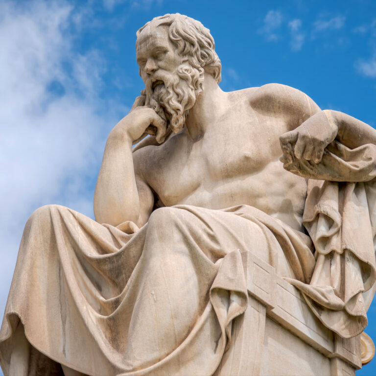 Close-up of a marble statue of a bearded male figure against a blue sky, highlighting the flowing hair and drapery